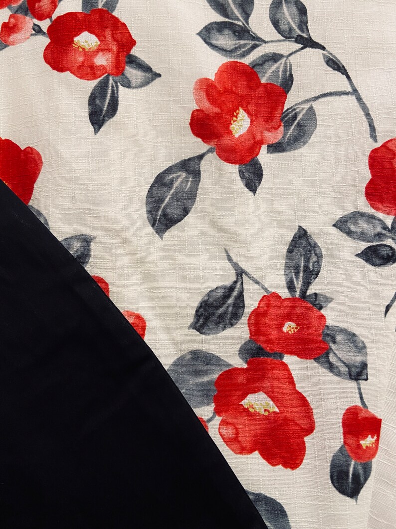Op de afbeelding: Een textielstof met een cr&egrave;mekleurige achtergrond en een herhalend patroon van felrode bloemen en donkergrijze bladeren. De stof heeft een zichtbare geweven textuur en wordt gedeeltelijk overlapt door een effen, glad, matzwart stoffen gedeelte.