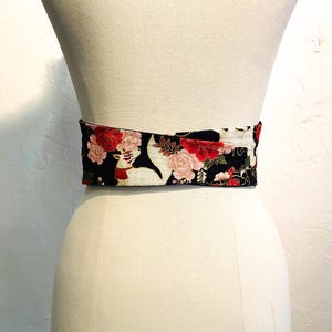 May include: A wide, decorative belt with a floral pattern. The belt features a black background with red and pink flowers and white accents. The belt is wrapped around a mannequin.