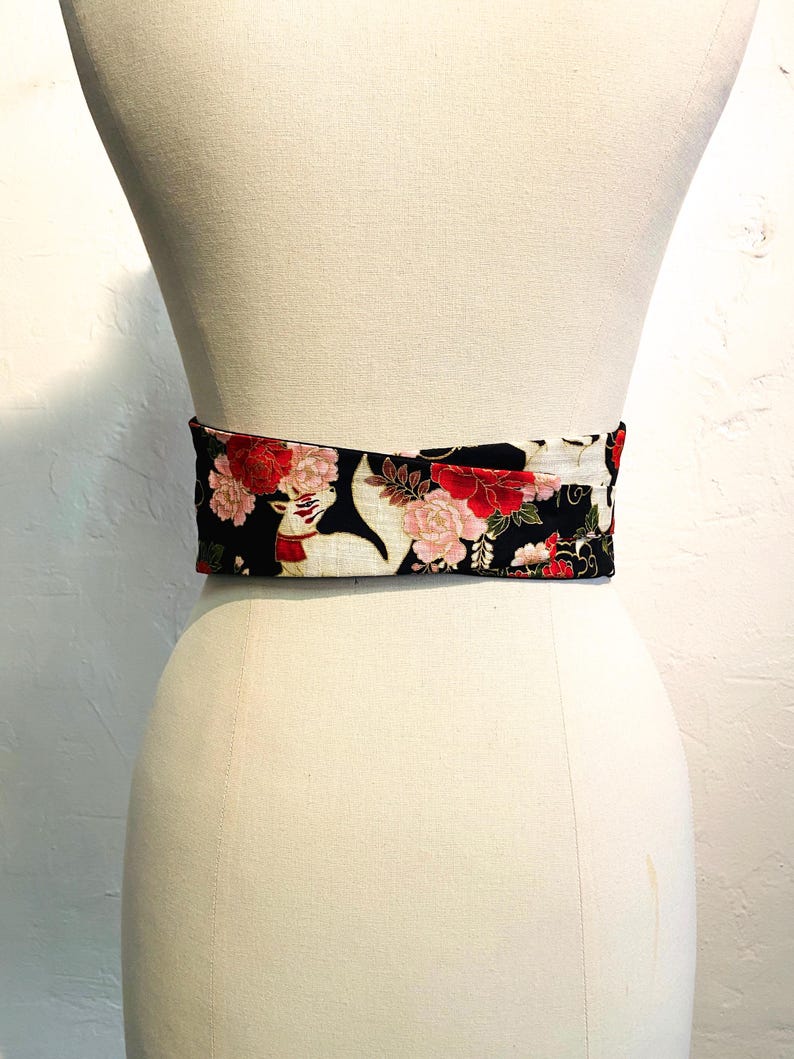 May include: A wide, decorative belt with a floral pattern. The belt features a black background with red and pink flowers and white accents. The belt is wrapped around a mannequin.