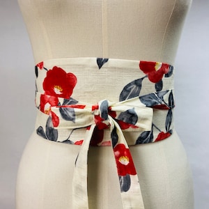Op de afbeelding: Een brede riem in obi-stijl van cr&egrave;mekleurige stof met een herhalend bloemmotief van felrode cameliabloesems en donkergrijze bladeren. De riem wordt om de taille gewikkeld en aan de voorzijde vastgemaakt met een gestrikte stoffen knoop.