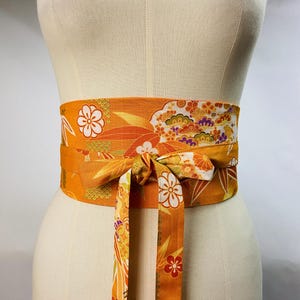 Puede incluir: Un cinturón ancho de tela naranja con un patrón floral. El cinturón está atado en un lazo en la parte delantera.