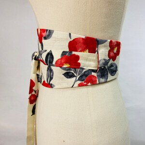 Op de afbeelding: Een brede obi-stijl wikkelriem met een cr&egrave;mekleurige stoffen basis en een bloemenpatroon van levendige rode cameliabloesems en donkergrijze bladeren. De riem heeft lange stoffen linten die om de taille worden gewikkeld en met een knoop vastzitten.