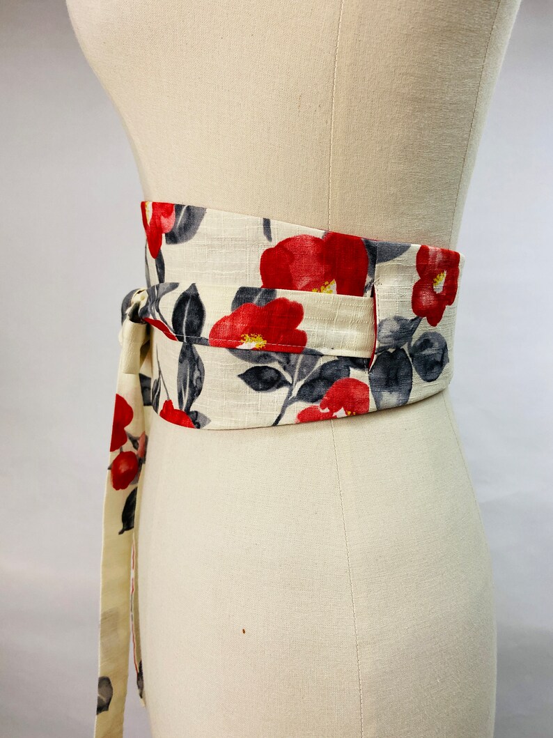 Op de afbeelding: Een brede obi-stijl wikkelriem met een cr&egrave;mekleurige stoffen basis en een bloemenpatroon van levendige rode cameliabloesems en donkergrijze bladeren. De riem heeft lange stoffen linten die om de taille worden gewikkeld en met een knoop vastzitten.