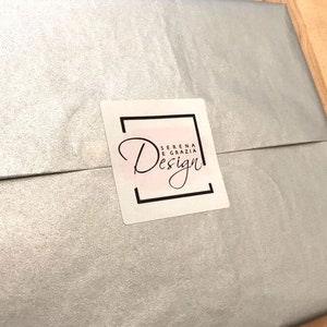 Op de afbeelding: Een witte envelop met een zilveren glimmende afwerking. Een wit vierkant label met een zwarte rand en de tekst "Serena e Grazia Design" bevindt zich aan de voorkant van de envelop.