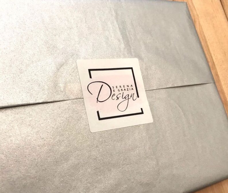Op de afbeelding: Een witte envelop met een zilveren glimmende afwerking. Een wit vierkant label met een zwarte rand en de tekst "Serena e Grazia Design" bevindt zich aan de voorkant van de envelop.