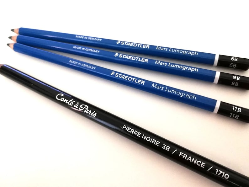 Op de afbeelding: Vier blauwe potloden met witte letters, gelabeld "Staedtler Mars Lumograph" en verschillende hardheidsgraden: 6B, 9B en 11B. Een zwart Conte &agrave; Paris potlood is ook afgebeeld, gelabeld "Pierre Noire 3B / France / 1710".