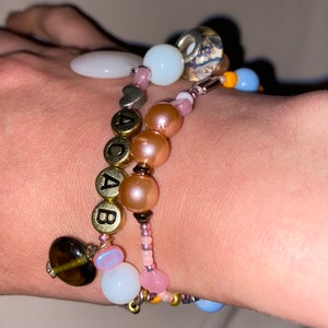 May include: A multi-layered bracelet with pink, white, and brown beads. The bracelet features a charm with the letters "ACAB" in gold.