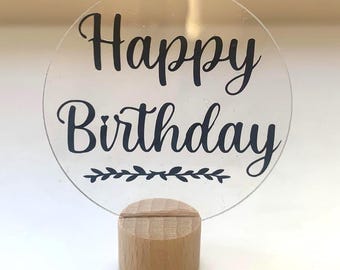以下が含まれることがあります： 黒い筆記体で「Happy Birthday」と書かれた透明な円形のサイン。サインは小さな天然木の台座で支えられています。「Birthday」という単語の下には、装飾的な葉のデザインがあります。