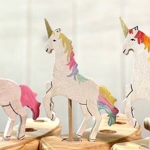 May include: Three wooden unicorn figurines with gold horns and colourful manes and tails. The unicorns are mounted on wooden dowels and set in a wooden base. The manes and tails are painted in shades of pink, yellow, and rainbow colours.