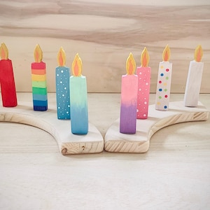 May include: A set of eight wooden candles with colorful painted designs. The candles are arranged on a wooden base in a semi-circle shape. The candles are painted in various colors, including red, blue, green, yellow, pink, and white.