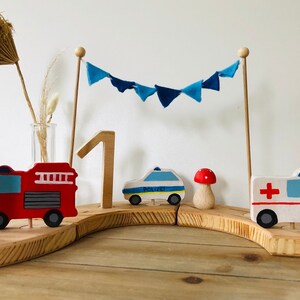 May include: A wooden first birthday cake topper with a red fire truck, a blue police car, a white ambulance, a mushroom, a rainbow candle, and a blue and white bunting.