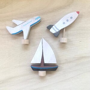 Könnte beinhalten: Drei Holzspielzeuge in Weiß, Blau und Braun bemalt. Die Spielzeuge sind ein Segelboot, ein Flugzeug und eine Rakete. Die Spielzeuge befinden sich alle auf einer Holzoberfläche.