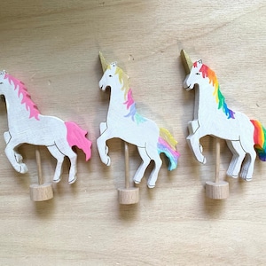 Pode incluir: Três figuras de unicórnios de madeira com corpos brancos, chifres dourados e crinas e caudas coloridas. Um tem crina e cauda rosa, outro cores pastel e o terceiro um arco-íris. Cada unicórnio é montado em uma base de madeira.