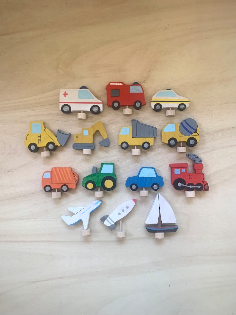 May include: A set of 15 wooden toy vehicles, including a fire truck, ambulance, police car, dump truck, cement mixer, excavator, tractor, airplane, rocket, sailboat, and train. The toys are painted in bright colors and have a whimsical design.