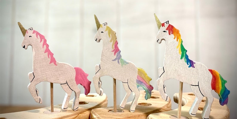 May include: Three wooden unicorn figurines with gold horns and colourful manes and tails. The unicorns are mounted on wooden dowels and set in a wooden base. The manes and tails are painted in shades of pink, yellow, and rainbow colours.