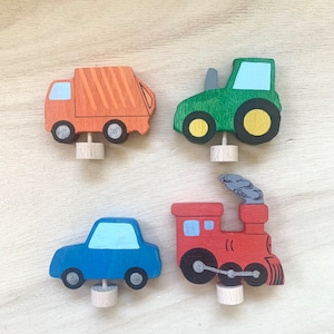 May include: Four wooden toy vehicles: a red train, a blue car, a green tractor, and an orange garbage truck. Each vehicle has a small wooden dowel for standing upright.