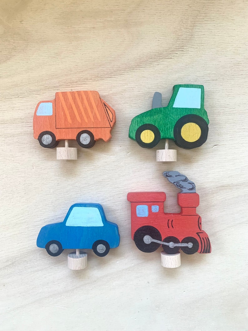 May include: Four wooden toy vehicles: a red train, a blue car, a green tractor, and an orange garbage truck. Each vehicle has a small wooden dowel for standing upright.
