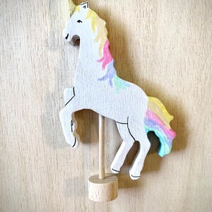May include: A wooden unicorn figurine with a gold horn and a rainbow mane and tail. The white unicorn is mounted on a wooden dowel and base. The unicorn is painted with pastel colours and stands on its hind legs.
