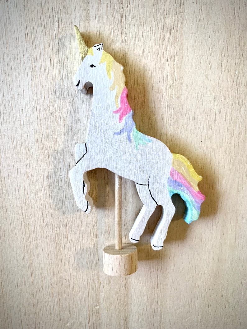 May include: A wooden unicorn figurine with a gold horn and a rainbow mane and tail. The white unicorn is mounted on a wooden dowel and base. The unicorn is painted with pastel colours and stands on its hind legs.