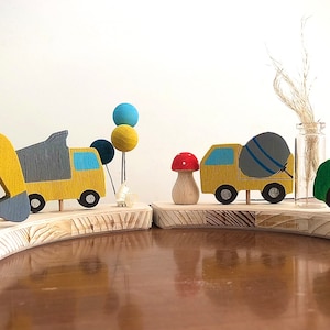 May include: A wooden toy set featuring a yellow excavator, a yellow dump truck with a gray bed, a yellow cement mixer truck with a gray drum, a green tractor, and a red mushroom. The toys are arranged on a wooden base with a light brown finish.