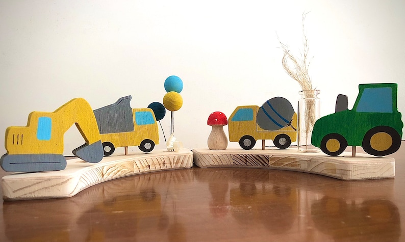 May include: A wooden toy set featuring a yellow excavator, a yellow dump truck with a gray bed, a yellow cement mixer truck with a gray drum, a green tractor, and a red mushroom. The toys are arranged on a wooden base with a light brown finish.