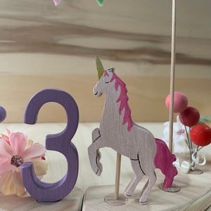 May include: A wooden unicorn cake topper with a pink mane and tail, a gold horn, and a wooden number 3 in purple. The topper is decorated with a garland of pastel-coloured felt flags and small pink and red decorative balls.