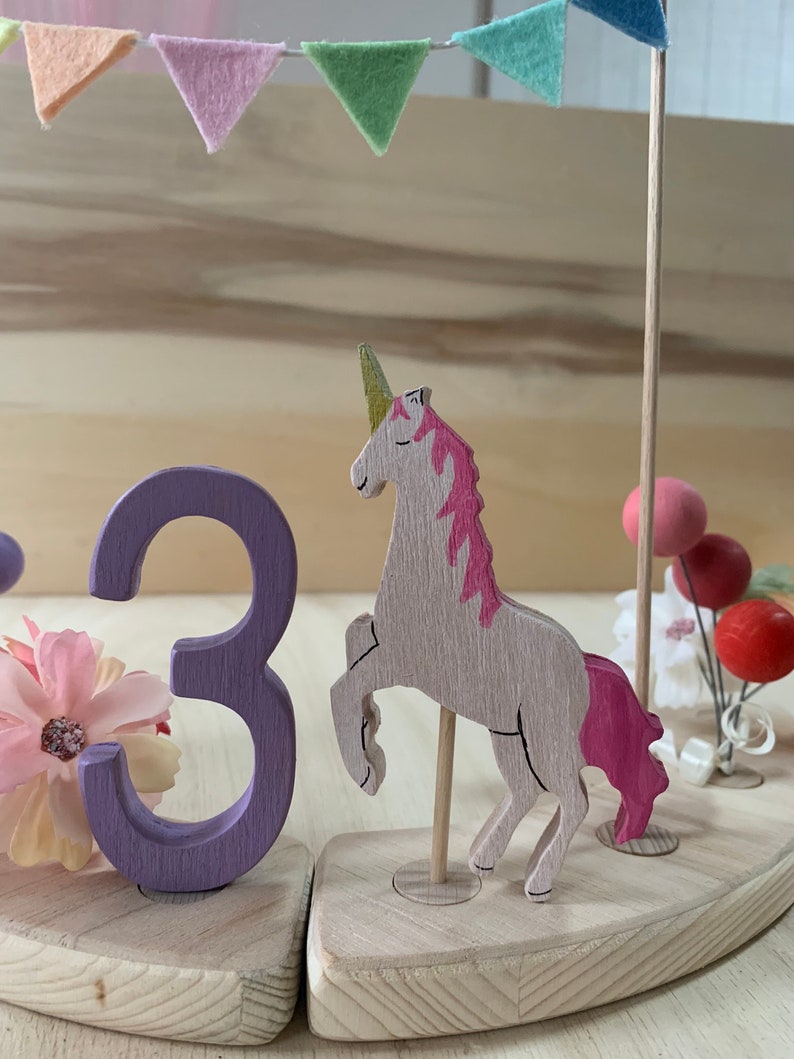 May include: A wooden unicorn cake topper with a pink mane and tail, a gold horn, and a wooden number 3 in purple. The topper is decorated with a garland of pastel-coloured felt flags and small pink and red decorative balls.