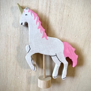 May include: A wooden unicorn figurine. It has a white body, pink mane and tail, and a gold horn. The unicorn is mounted on a wooden stand. Black lines outline the features. A whimsical decorative item.