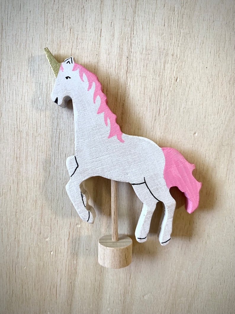 May include: A wooden unicorn figurine. It has a white body, pink mane and tail, and a gold horn. The unicorn is mounted on a wooden stand. Black lines outline the features. A whimsical decorative item.