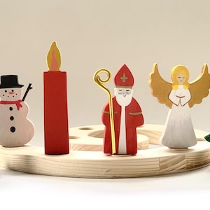 May include: A wooden circular base holds six painted figures: a green tree, a white snowman with a black hat, a red candle, a figure in red robes with a gold staff, a white angel with gold wings, and a green tree topped with a gold star.