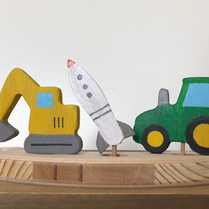 May include: A wooden toy set with a yellow excavator, a green tractor, and a white rocket. The toys are painted with simple details and are arranged on a wooden base.