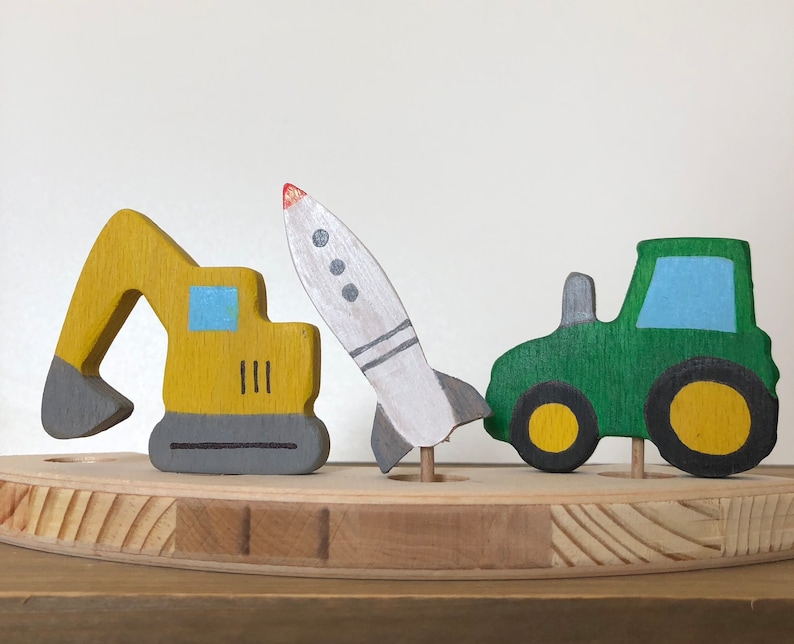 May include: A wooden toy set with a yellow excavator, a green tractor, and a white rocket. The toys are painted with simple details and are arranged on a wooden base.