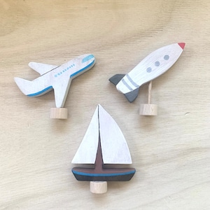 May include: Three wooden toys painted in white, blue, red, and black. The toys are a sailboat, an airplane, and a rocket. The toys are all on a wooden surface.