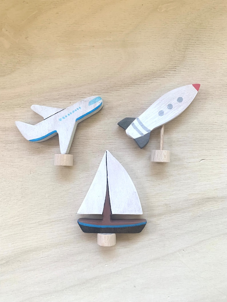 May include: Three wooden toys painted in white, blue, red, and black. The toys are a sailboat, an airplane, and a rocket. The toys are all on a wooden surface.