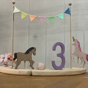 May include: Wooden birthday cake topper featuring a horse, unicorn, and the number 3. The topper includes a banner with pastel flags and a small vase of pink flowers. The base is made of light-coloured wood.