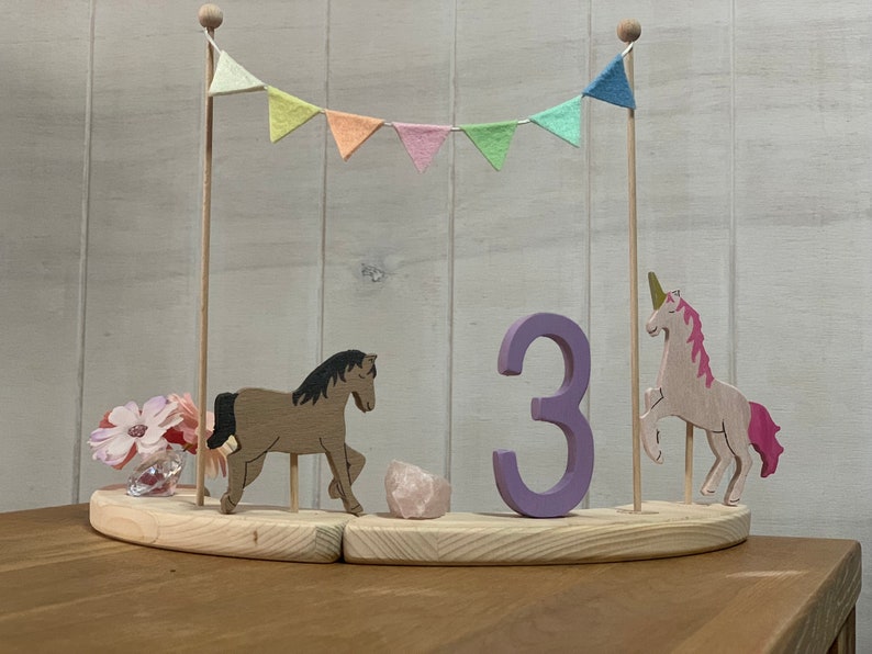 May include: Wooden birthday cake topper featuring a horse, unicorn, and the number 3. The topper includes a banner with pastel flags and a small vase of pink flowers. The base is made of light-coloured wood.
