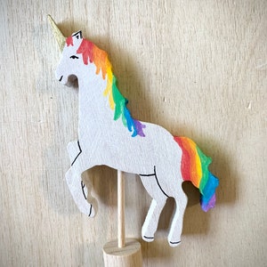 May include: A wooden unicorn figurine. It has a white body, a rainbow mane and tail, and a gold horn. The unicorn is mounted on a wooden stand. The mane and tail display a vibrant spectrum of colours.