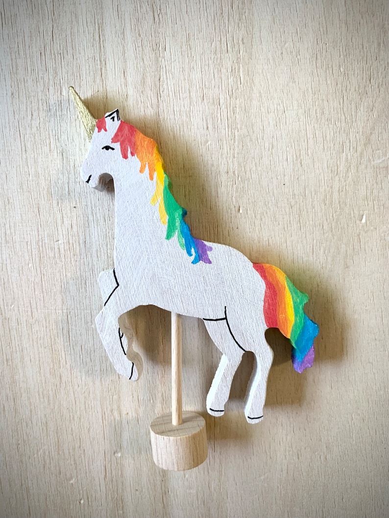 May include: A wooden unicorn figurine. It has a white body, a rainbow mane and tail, and a gold horn. The unicorn is mounted on a wooden stand. The mane and tail display a vibrant spectrum of colours.