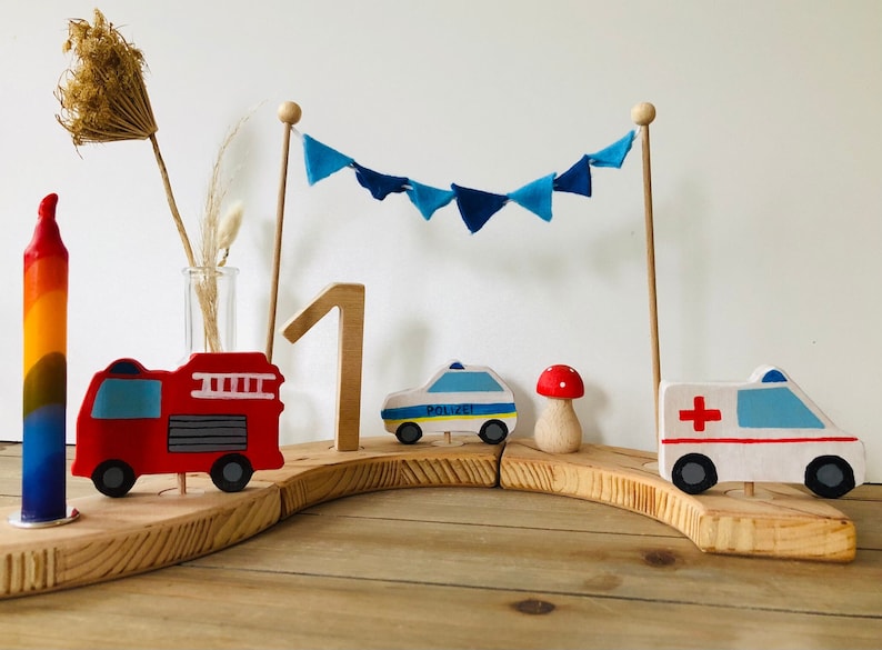 May include: A wooden first birthday cake topper with a red fire truck, a blue police car, a white ambulance, a mushroom, a rainbow candle, and a blue and white bunting.
