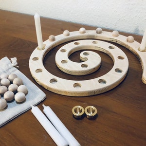 Könnte beinhalten: Ein Adventskalender aus Holz in Form einer Spirale mit 24 nummerierten Löchern zum Platzieren kleiner Holzperlen. Der Kalender hat zwei Kerzenhalter und wird mit zwei weißen Kerzen und zwei Messingkerzenhaltern geliefert.