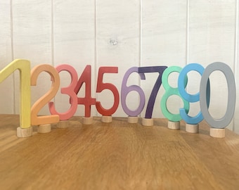 May include: Wooden numbers 1 to 10 painted in rainbow colours. The numbers are standing on small wooden bases.