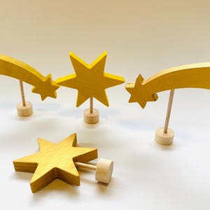 Könnte beinhalten: Vier Holzstern-Dekorationen in einem warmen Gelbton. Zwei sind Sternschnuppen und zwei sind klassische fünfzackige Sterne. Jeder Stern ist auf einem kleinen Holzdübel und Sockel montiert, was eine skurrile, handgefertigte Ästhetik erzeugt.