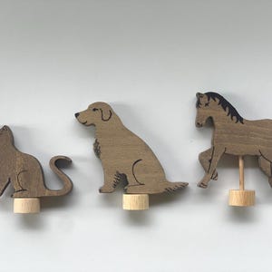 Könnte beinhalten: Drei Holztiere: eine Katze, ein Hund und ein Pferd. Die Katze sitzt, der Hund sitzt und das Pferd bewegt sich. Jede Figur ist braun mit schwarzen Akzenten und auf einem kleinen Holzsockel montiert.