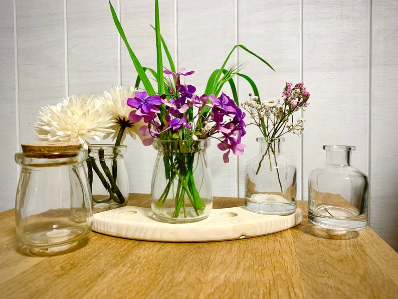 以下が含まれることがあります： 透明なガラスの花瓶4つに、さまざまな種類の花が活けられています。花瓶は木製の台の上に置かれています。花には、白い菊、紫と白の野の花、小さな白い花があります。