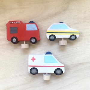 May include: Three wooden toy vehicles: a red fire truck, a white and yellow police car, and a white ambulance with a red cross. The vehicles have black wheels and are painted with details.