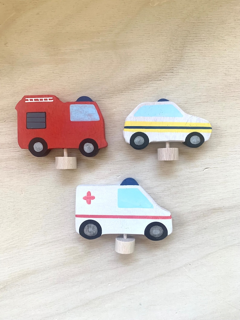 May include: Three wooden toy vehicles: a red fire truck, a white and yellow police car, and a white ambulance with a red cross. The vehicles have black wheels and are painted with details.
