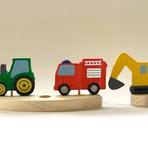 Pode incluir: Conjunto de brinquedos de madeira com um trator verde, um carro de bombeiros vermelho e uma escavadeira amarela. Os veículos são montados em uma base de madeira clara. Brinquedo infantil.