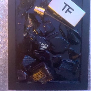 May include: A black framed display featuring a perfume bottle with a white and black label with the letters "TF". The display includes black shards and the text "TOM FORD NOIR EXTREME". The perfume bottle has a gold cap.