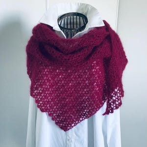 Peut inclure: Un châle triangulaire magenta profond présentant un motif de crochet ouvert et aéré avec un aspect texturé et duveteux. Le châle est drapé sur une chemise blanche à col boutonné, mettant en valeur le contraste entre le fil rouge foncé et le tissu.