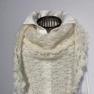 Peut inclure: Un châle triangulaire crème avec un motif ajouré délicat. Le châle a un bord frangé et est drapé sur une chemise blanche à col. La texture semble douce et aérée, suggérant un matériau léger.